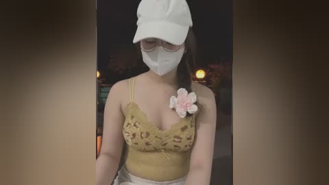 Media: Video of a young Asian woman in a white cap, mask, and glasses, wearing a yellow lace camisole with a pink flower brooch, standing in a dimly lit room.