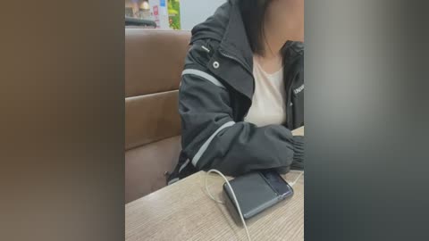 Media: Video of a woman in a black jacket and white top, seated at a wooden table with a smartphone and headphones. Background shows a brown leather seat and blurred objects.