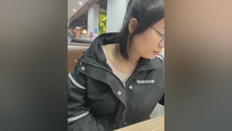 Media: Video of a young Asian woman with straight black hair, wearing glasses and a black jacket with white stripes, sitting at a table in a dimly lit restaurant.