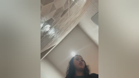 Media: A video of a woman with long, dark hair and a light skin tone, captured from below, showing her upper body and face. She stands in an indoor setting with a geometric, glass ceiling and white walls.