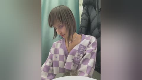 Media: Video of a young woman with short, straight brown hair, wearing a purple and white checkered sweater, sitting in a dark, padded chair in a dimly lit room with teal curtains in the background.