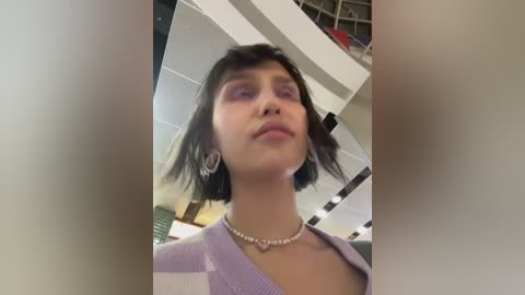 Media: Video of a young woman with short, dark hair, wearing a lavender cardigan, silver hoop earrings, and a pearl necklace, standing in a modern indoor setting with white walls and a metal structure.