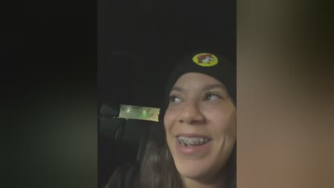 Media: Video of a young woman with braces, wearing a black beanie, smiling broadly. The background is dark, creating a contrast that highlights her happy expression.