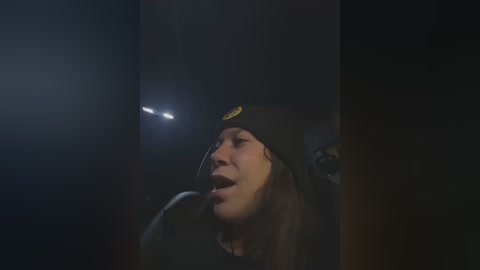 Media: Video of a woman with long brown hair, wearing a black beanie with a yellow logo, singing into a microphone, in a dimly lit, shadowy room with a small spotlight on her face.