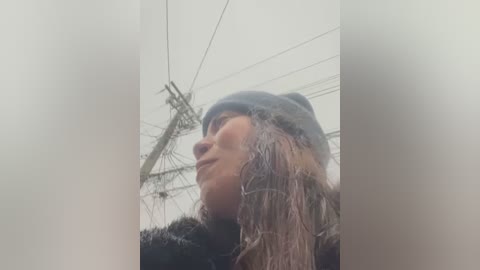 Media: A video of a woman with long, wet hair, wearing a black beanie and coat, standing in a foggy, industrial setting with a crane and cables in the background.