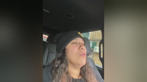 Media: A video of a young woman with long, wavy brown hair, wearing a black beanie and dark jacket, leaning back with eyes closed, inside a car with a green and brown patterned wall visible through the window.