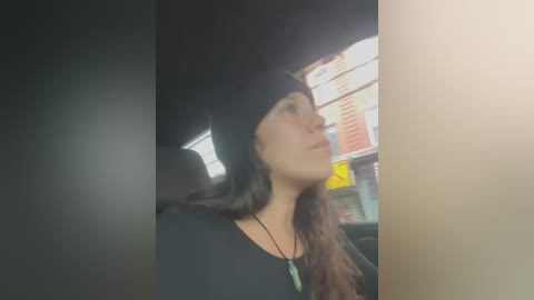 Media: Video of a young woman with long dark hair, wearing a black beanie and black top, sitting in a dimly lit car, with a blurred storefront in the background.