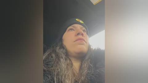 Media: A video of a woman with fair skin, wearing a black beanie with a circular emblem and a dark, furry coat, gazing upwards. The background is dark and slightly blurry, with a bright, circular light source on the right.