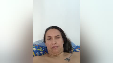 Media: A video of a middle-aged woman with medium skin tone and long black hair, sitting shirtless on a colorful, patterned pillow, against a plain white wall, appearing contemplative.