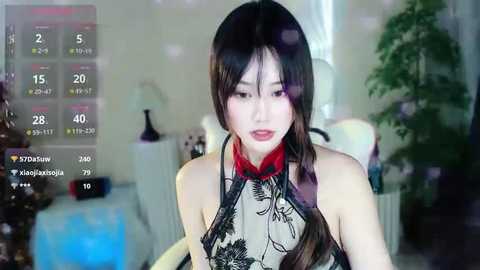 Media: Video of a young Asian woman with long black hair, wearing a traditional black and red cheongsam, seated in a dimly lit room with a potted plant in the background.