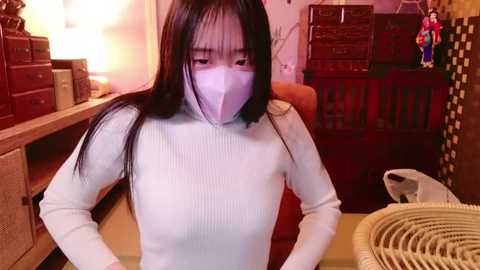 Media: Video of an Asian woman with long black hair wearing a white ribbed sweater, face mask, and a beige backpack, sitting indoors with wooden furniture and a wicker basket in the background.