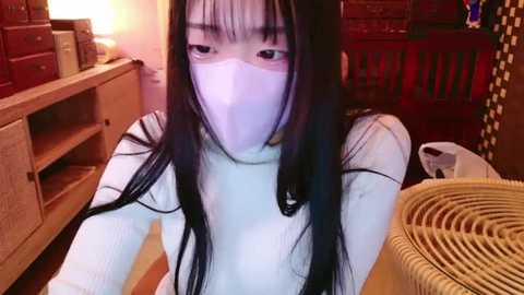 Media: A video of an East Asian woman with long black hair, wearing a white turtleneck and white mask, sitting in a cozy room with wooden furniture, including a red crib and wicker basket.