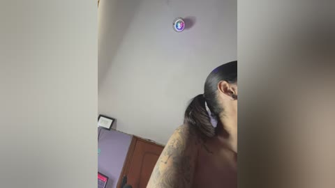 Media: A video of a shirtless man with a beard, tattoos, and long black hair tied back, partially seen in profile, standing indoors next to a wall-mounted circular light fixture.