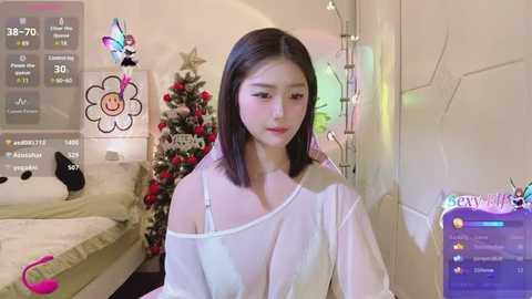 Media: Video of an East Asian woman with straight black hair, wearing a white off-shoulder top, in a cozy, decorated room with a Christmas tree, virtual butterfly overlay, and a \"Caty Cat\" game screen.