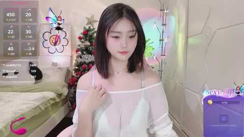 Media: Video of a young Asian woman with straight, shoulder-length black hair, wearing a white, off-the-shoulder lace dress, standing indoors next to a decorated Christmas tree.