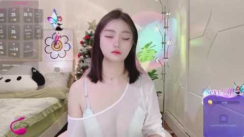 Media: A video of an Asian woman with medium-length black hair, fair skin, and a slim physique, wearing a sheer white top, in a cozy, decorated bedroom with a digital calendar, plush toys, and a rainbow effect.