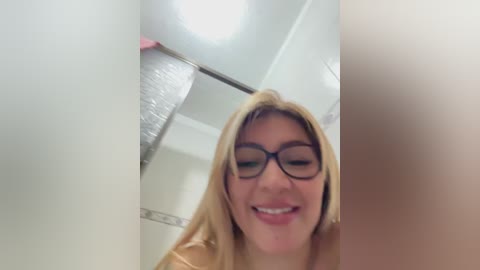 Media: A video of a smiling, light-skinned woman with long blonde hair and black-framed glasses, standing in a bathroom with white walls, a shower curtain, and a visible ceiling light.