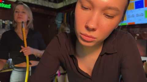 Media: Video of a young woman with short brown hair and a nose ring, wearing a dark top, looking contemplative in a dimly-lit bar. Another woman with blonde hair and glasses stands behind her, holding a microphone.