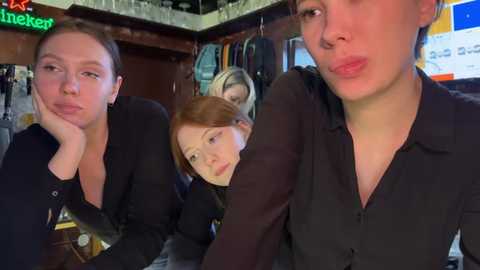 Media: Video of three women in a dimly lit bar, two with red hair, one blonde, all wearing black tops. Background includes bar shelves, neon \"Heineken\" sign, and blurred patrons.