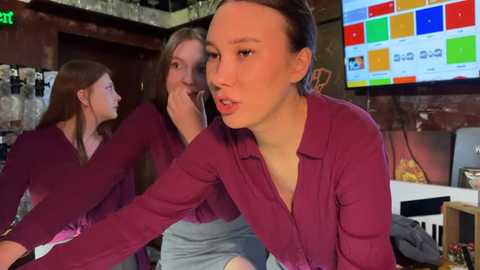 Media: Video of three young women playing video games in a dimly lit room with a TV screen showing colorful game graphics. They are wearing matching maroon shirts and gray pants.