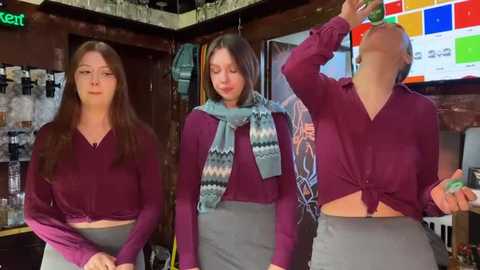 Media: Video of three young women with long hair, wearing maroon sweaters and gray skirts, holding green drinks, standing in a cozy, dimly-lit bar with colorful posters and a neon sign.