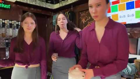 Media: Video of three young women in burgundy tops and grey skirts, working behind a bar with colorful beer signs in the background.