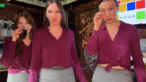 Media: Video of three young women in a dimly lit bar, all wearing maroon tops and gray pants, chatting and smiling. Background features colorful chalkboard art and bar paraphernalia.