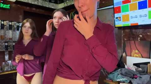 Media: Video of a young woman in a red shirt, tied at the waist, and maroon underwear, standing in a dimly-lit bar. Background shows two other women in similar attire, with one holding a phone.