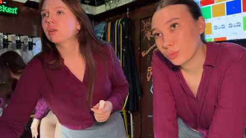 Media: Video of two young women with light skin and brown hair, wearing maroon tops, standing in a cluttered, dimly-lit room with clothes hanging on racks and a TV screen displaying colorful graphics.