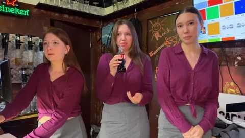 Media: Video of three young women in a bar, holding a glass of beer. They wear matching maroon tops and grey skirts, standing near a colorful, abstract wall painting.