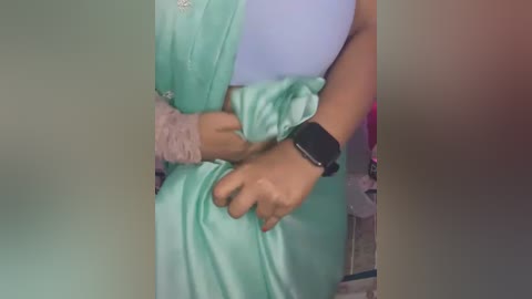 Media: Video of a person in a light green saree with a pink beaded wristband, wearing a black smartwatch. The background is blurred, featuring a soft gradient of beige and pink tones.