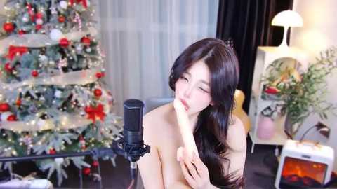 Media: Video of an Asian woman with long black hair, wearing a crown, seductively licking a microphone in a festive room with a decorated Christmas tree and lit candles.