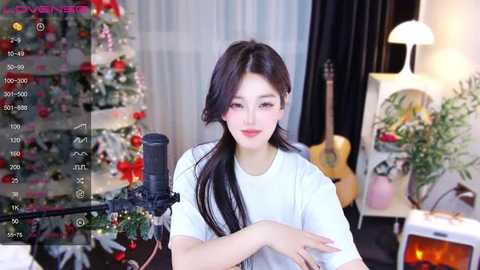 Media: A video of an Asian woman with long black hair, wearing a white shirt, sitting in front of a microphone in a cozy, Christmas-decorated room with a guitar and flowers.