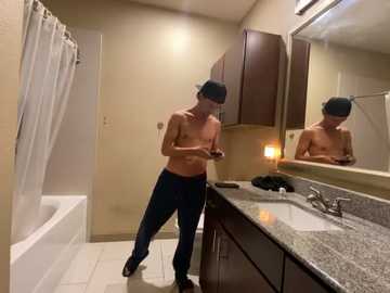 Media: Video of a shirtless man in dark pants, wearing a black beanie, using a smartphone, standing in a beige-tiled bathroom with a granite countertop, a mirror, and a bathtub.