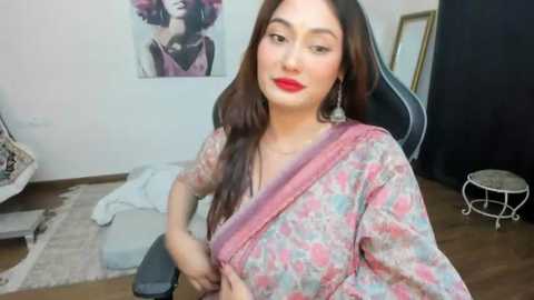 Media: Video of a smiling South Asian woman with long brown hair, wearing a pink floral saree, red lipstick, sitting on a gaming chair, in a modern, minimalist room with a framed artwork and a white stool.