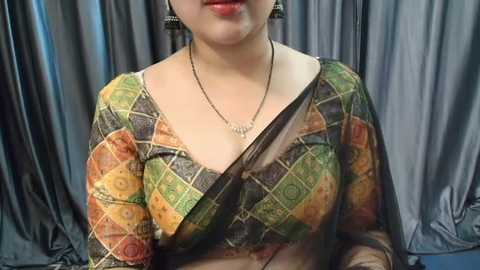 Media: Video of a light-skinned woman wearing a vibrant, patchwork sari with geometric patterns in green, orange, and brown. She has a delicate necklace and earrings. Dark grey curtains form the background.