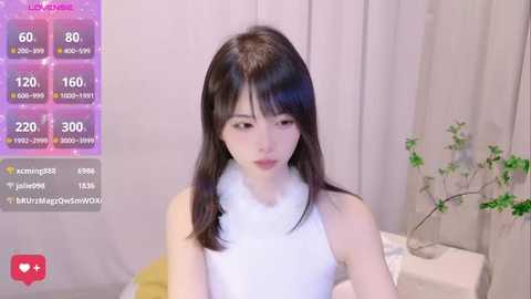 Media: Video of an East Asian woman with straight, dark hair and fair skin, wearing a white sleeveless top with a fluffy collar, in a cozy indoor setting.