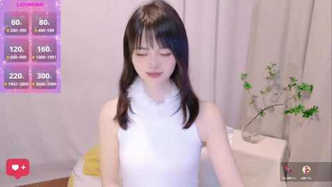 Media: Video of a young Asian woman with long black hair, wearing a white sleeveless dress, standing in a minimalist room with light-colored curtains, a potted plant, and a pink heart overlay.