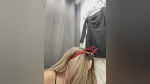 Media: Video of a blonde woman with straight hair, wearing a red headband, bending over in a dimly lit, narrow hallway with gray walls and black coats hanging on hooks.