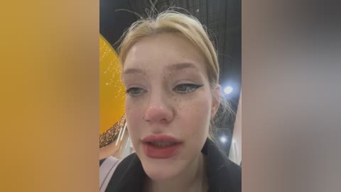 Media: Video of a young Caucasian woman with fair skin, freckles, and blonde hair. She has bold black eyeliner and red lipstick, wearing a black top. Background features a blurred indoor setting with a yellow and orange balloon.