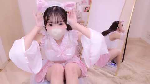 Media: Video of an Asian woman with light skin and long black hair, wearing a pink frilly maid outfit, with a pink bow, and a white mask, sitting in front of a mirror.