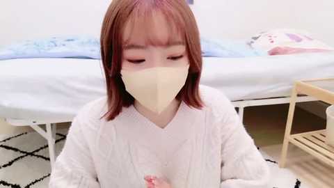 Media: A video of a young Asian woman with fair skin, straight auburn hair, and a beige face mask, wearing a white bathrobe, sitting on a patterned rug in a minimalist bedroom with a bed and wooden stool.