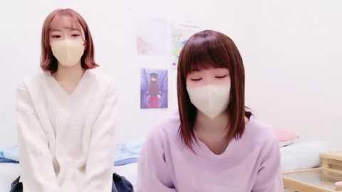 Media: Video of two young East Asian women with straight brown hair, wearing white face masks and light-colored robes, sitting in a minimalist bedroom with white walls and colorful posters.