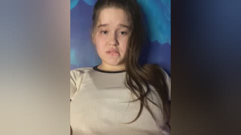 Media: Video of a young Caucasian girl with light skin and long brown hair, wearing a beige ribbed shirt, against a blue and purple abstract background. She has a solemn expression.