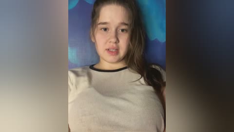 Media: Video of a young Caucasian woman with fair skin and straight brown hair tied back, wearing a fitted, ribbed, light grey top that accentuates her large breasts. She has a neutral expression and stands against a blue and purple abstract wall.