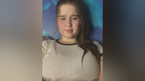 Media: Video of a young Caucasian woman with fair skin, long brown hair, and a chubby physique, wearing a beige ribbed t-shirt, standing against a blue and green abstract background.