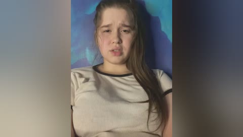 Media: Video of a young Caucasian woman with fair skin, long brown hair, wearing a light gray ribbed top, standing against a blue and purple backdrop, looking sad with tears in her eyes.