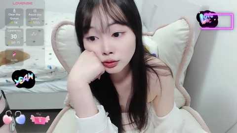 Video of an Asian woman with long black hair, wearing a white off-shoulder sweater, lying on a bed with pillows, looking tired and sad.