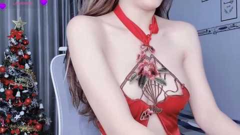 Media: A video of an Asian woman with long brown hair, wearing a red lace bra with floral embroidery, sitting next to a decorated Christmas tree in a cozy, festive room.