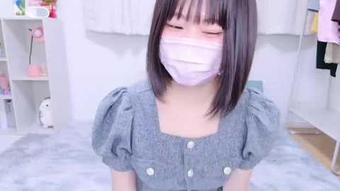 Media: Video of a young Asian woman with straight black hair and fair skin, wearing a gray blouse, pink face mask, and blush, in a white-walled room with a white floor, a shelf with toys, and a pink towel.